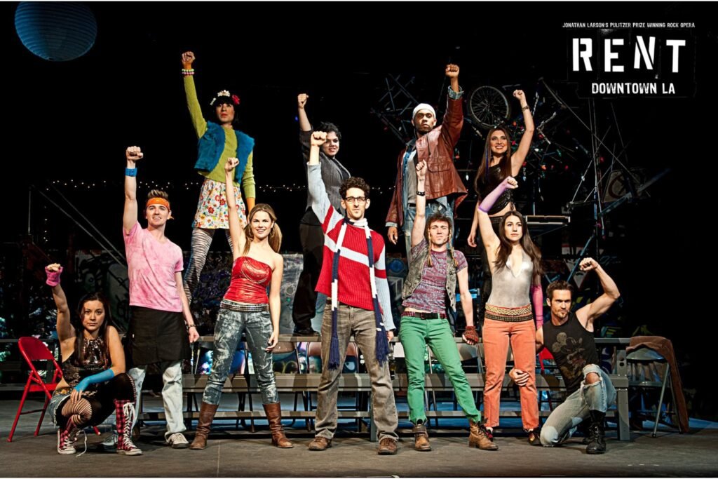 Still shot of theatre production RENT featuring full cast in costume including iconic red and blue knit sweater