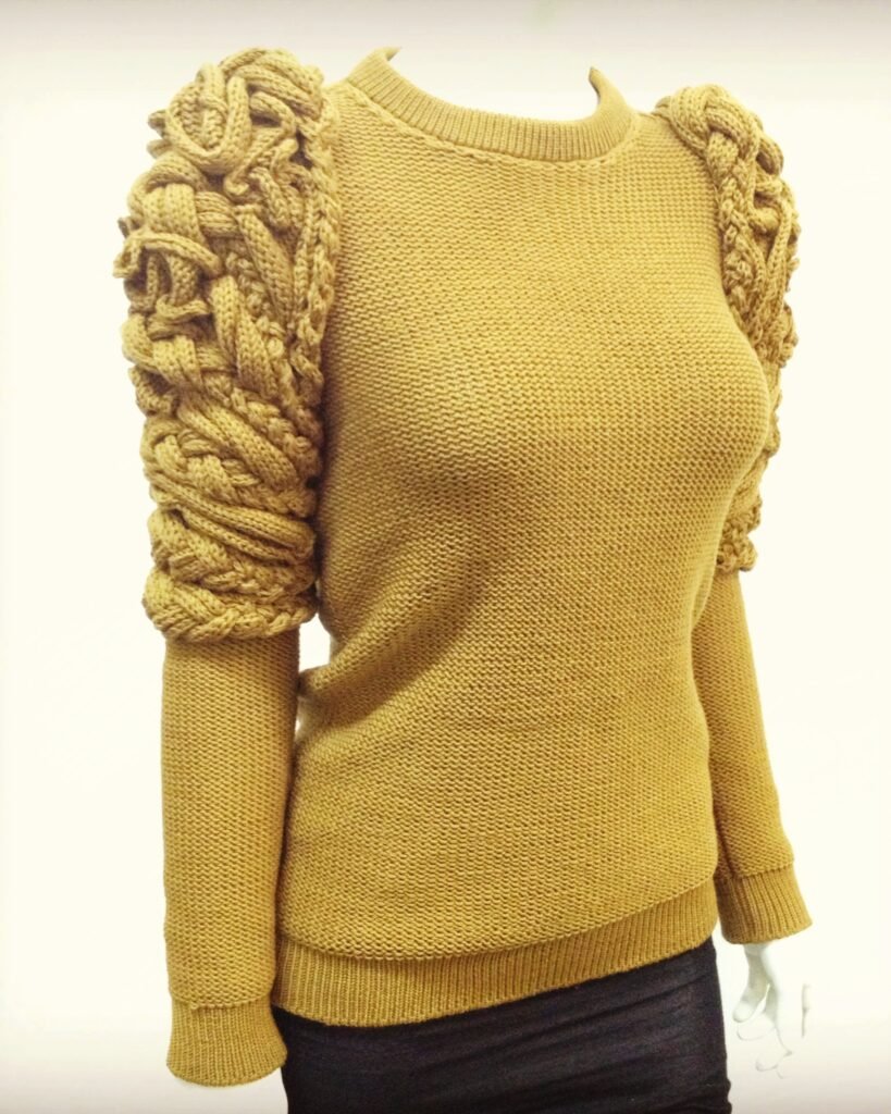 Camel colored knit sweater featuring sculptural braided elements on shoulders and upper arms, worn on a mannequin