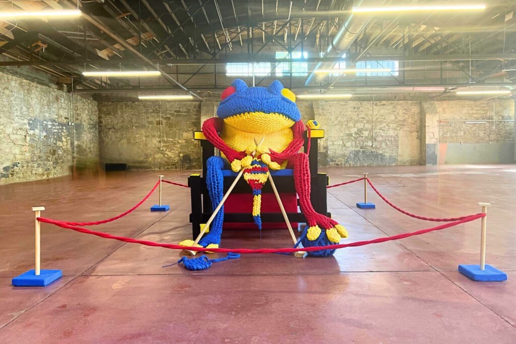 FROG - A Fiber Situation installation showing large-scale sculpture of a frog knitting in a chair, installed in a large warehouse gallery