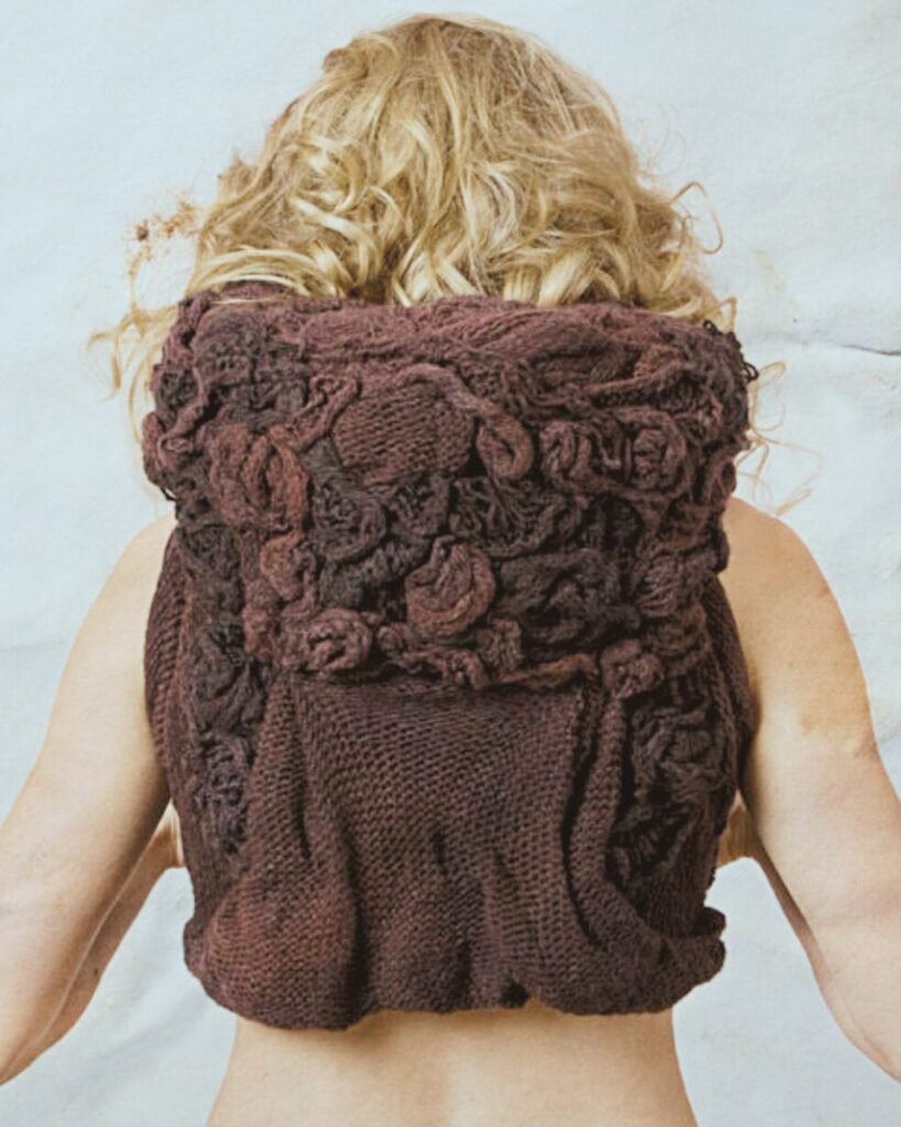 Back of model seen wearing a purple knit vest featuring sculptural elements that mimic flowers